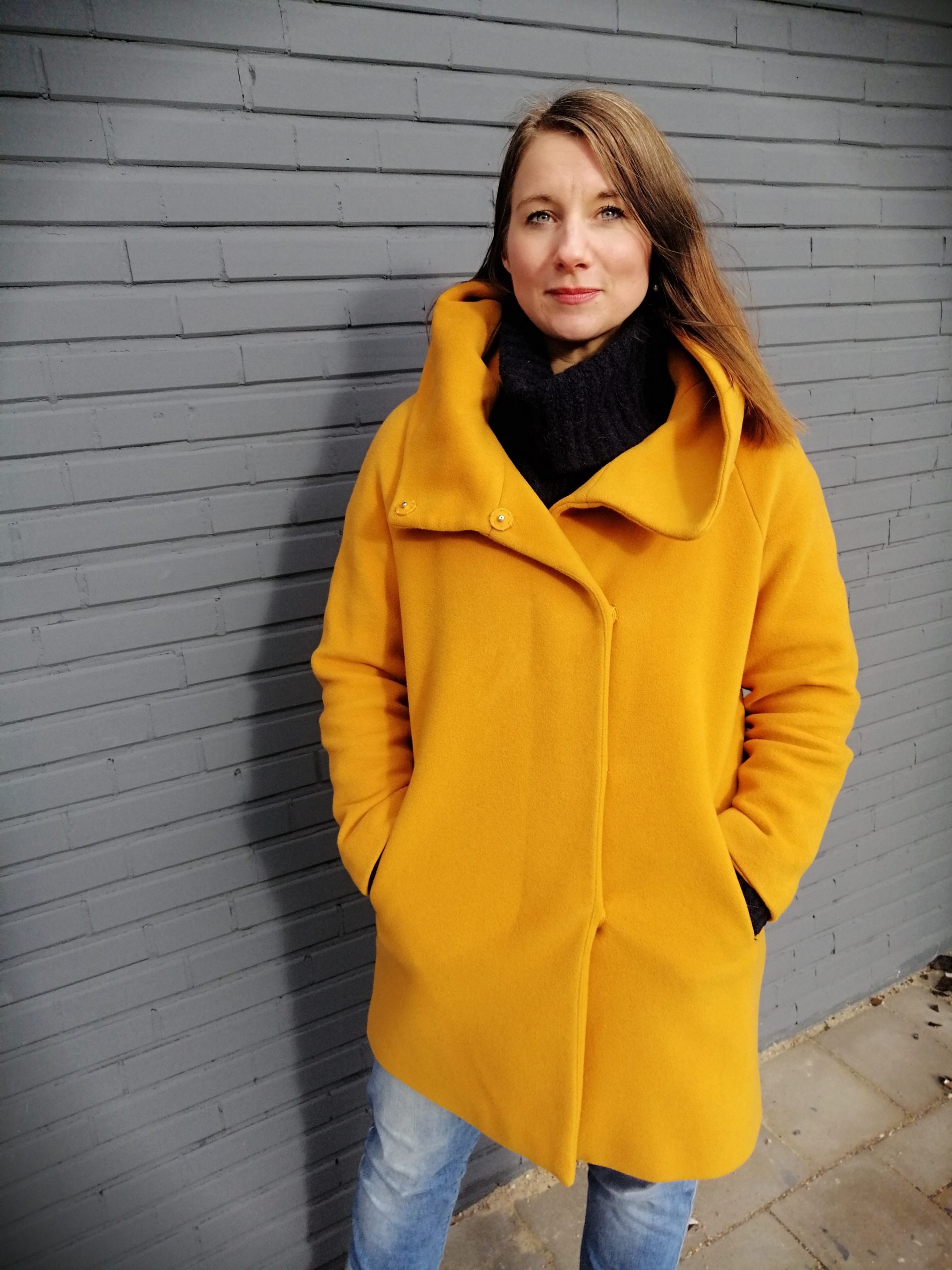 Mirja steht mit einem gelben Mantel vor einer grauen Wand.