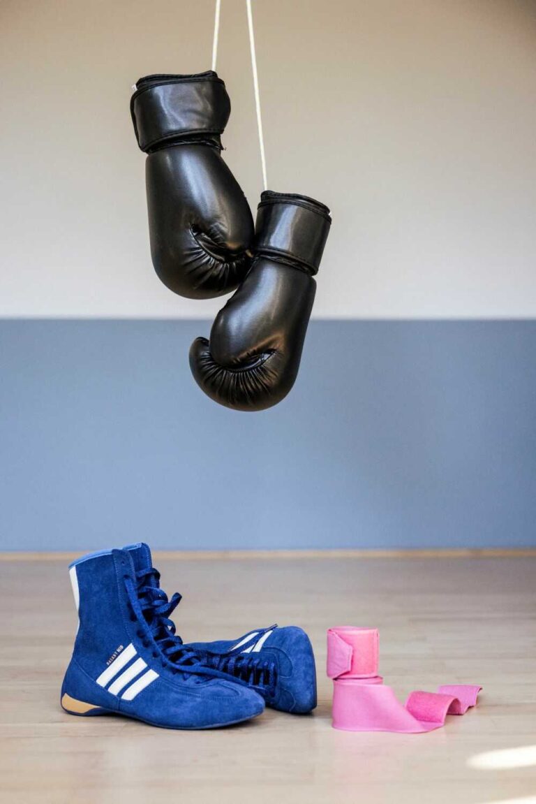 Schwarze Boxhandschuhe hängen an weißer Schnur, darunter blaue Boxschuhe und rosa Handbandagen